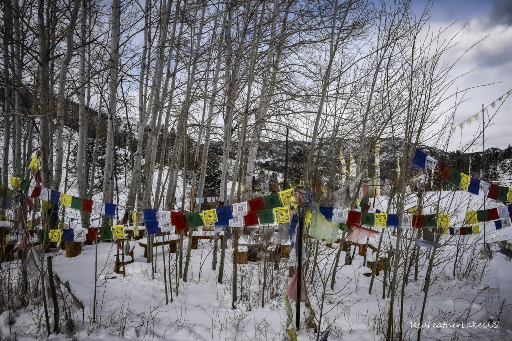 Colorful Buddhist prayer flags in snowy aspen forest with stormy skies and mountains in the backdrop at Drala Mountain Center, Red Feather Lakes Colorado