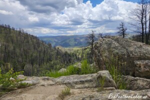 The same Cameron Peak Fire burn scar from Boy Scout Road (County Road 69) in Northwest Larimer County, Colorado in July 2023, three years after the fire — standing dead trees above a granite outcrop, with grass and young conifers greening the valley below under summer storm clouds