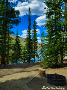 A Chambers Lake Campground site on a bluff, a fire ring in the foreground and the blue lake glowing through tall spruce, in Colorado's Poudre Canyon