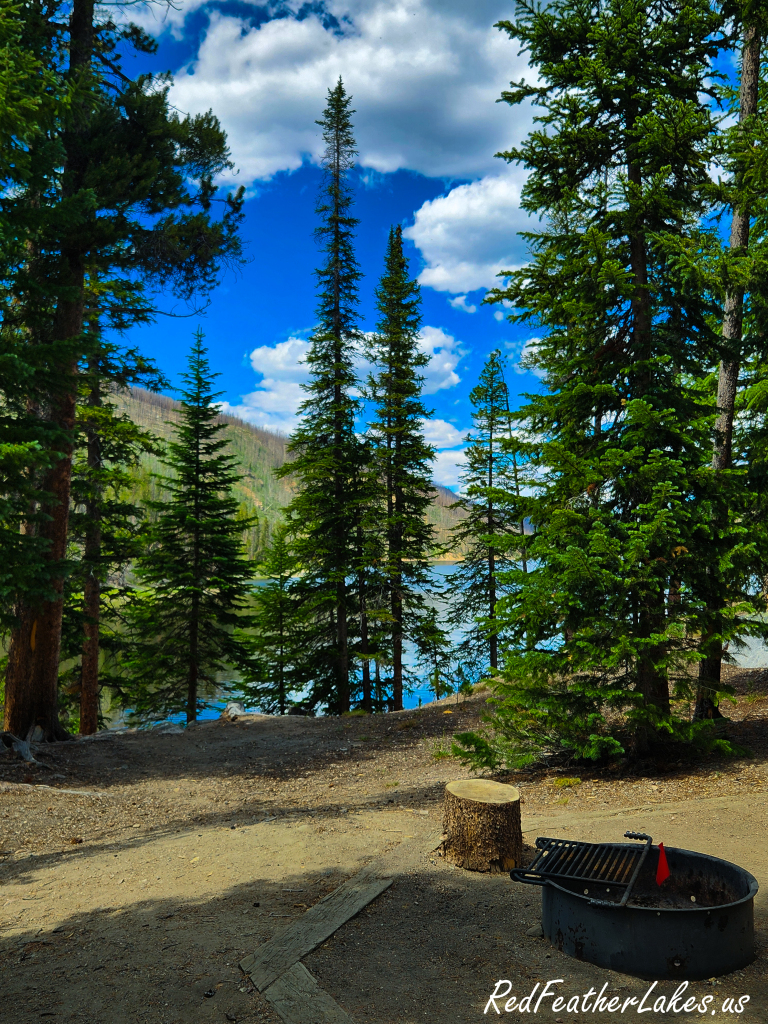 A Chambers Lake Campground site on a bluff, a fire ring in the foreground and the blue lake glowing through tall spruce, in Colorado's Poudre Canyon